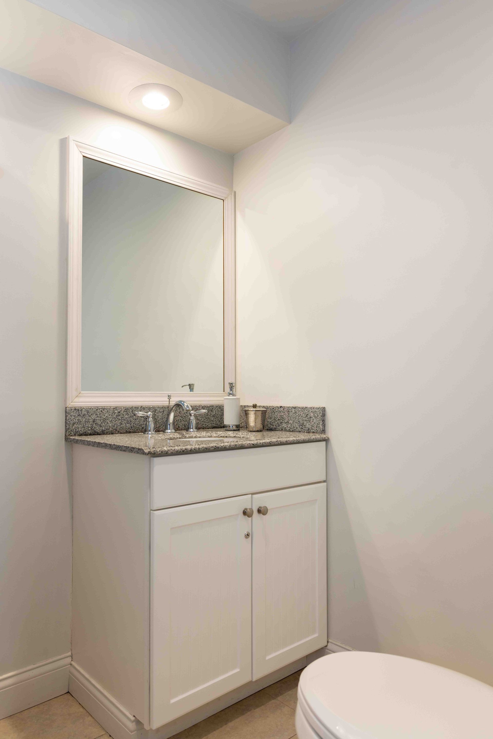 Small bathroom vanity with granite countertop, mirror, and white cabinet beside a toilet.