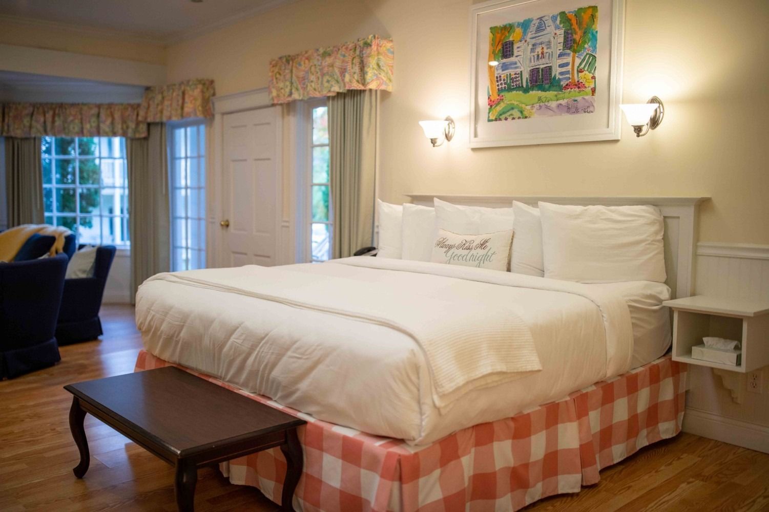 Cozy bedroom with a large white bed, checkered bed skirt, wooden table, and bright windows