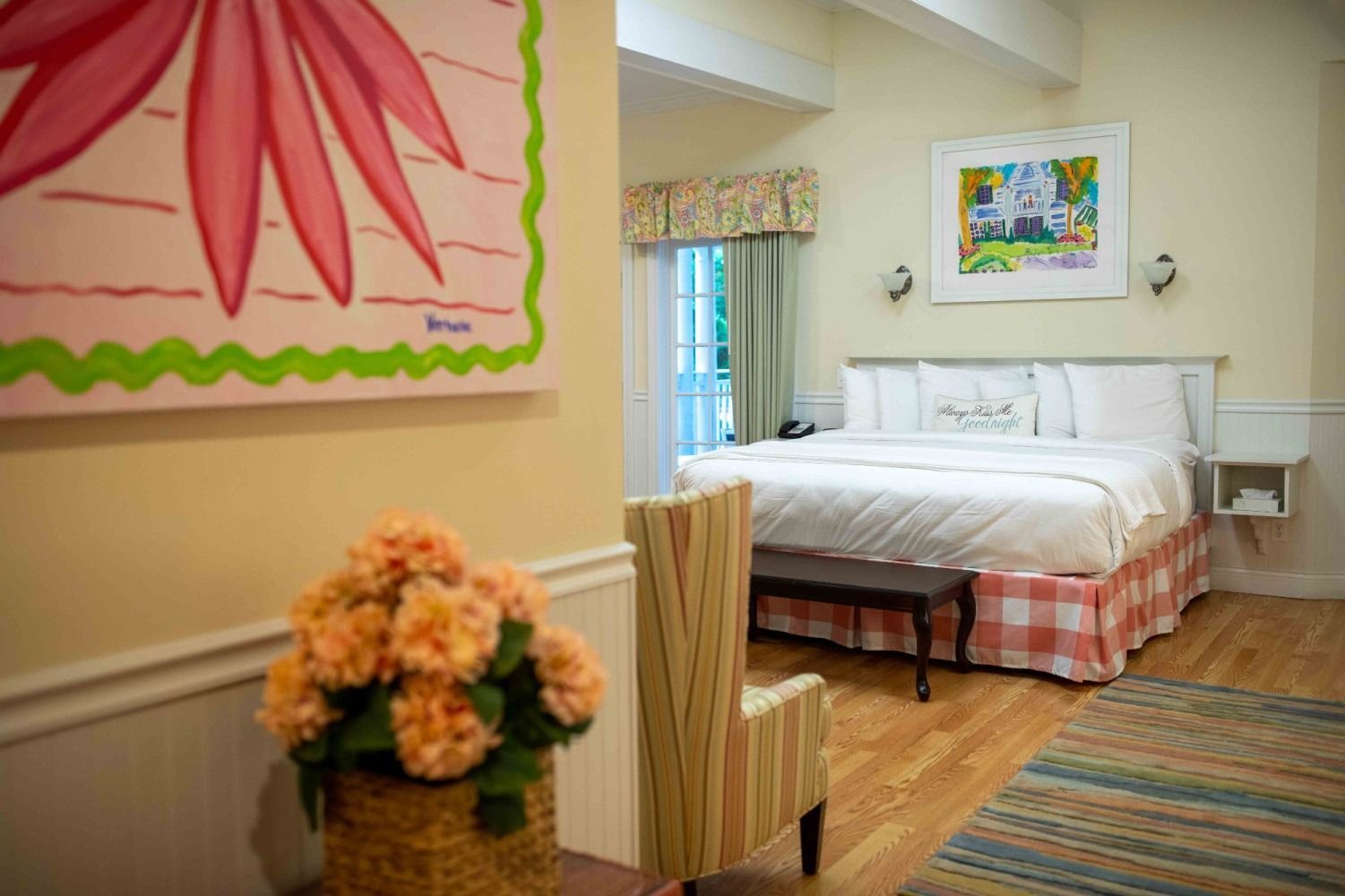 Bright bedroom with white bed, colorful wall art, striped rug, and floral decor in a cozy home interior.