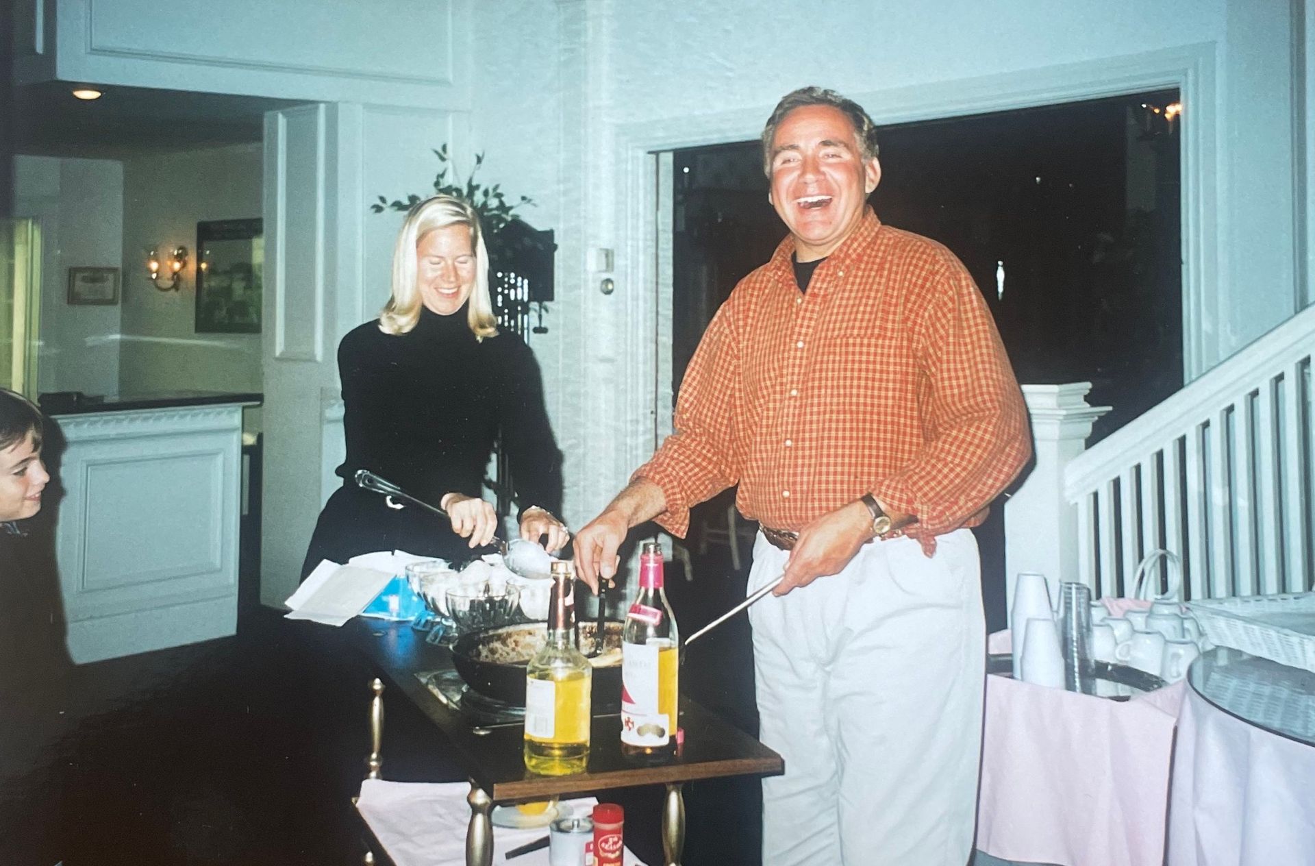 Two people smiling beside a table with drinks at a home gathering, in a bright kitchen area.