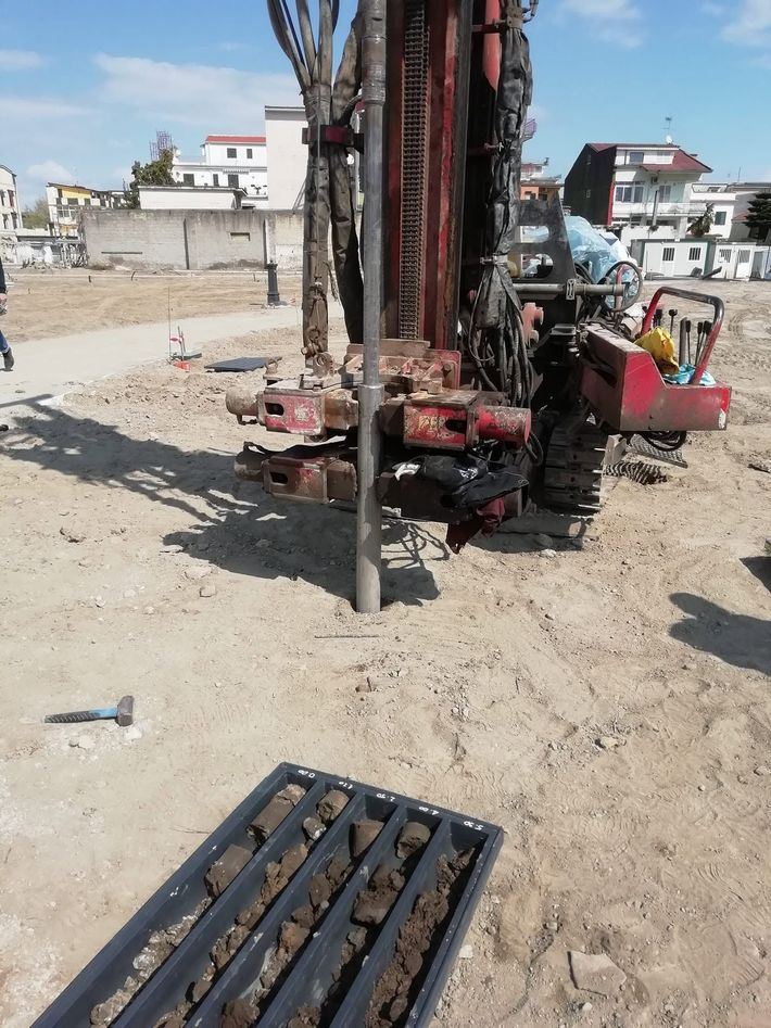 Impianto di perforazione in un cantiere sabbioso, con edifici sullo sfondo.