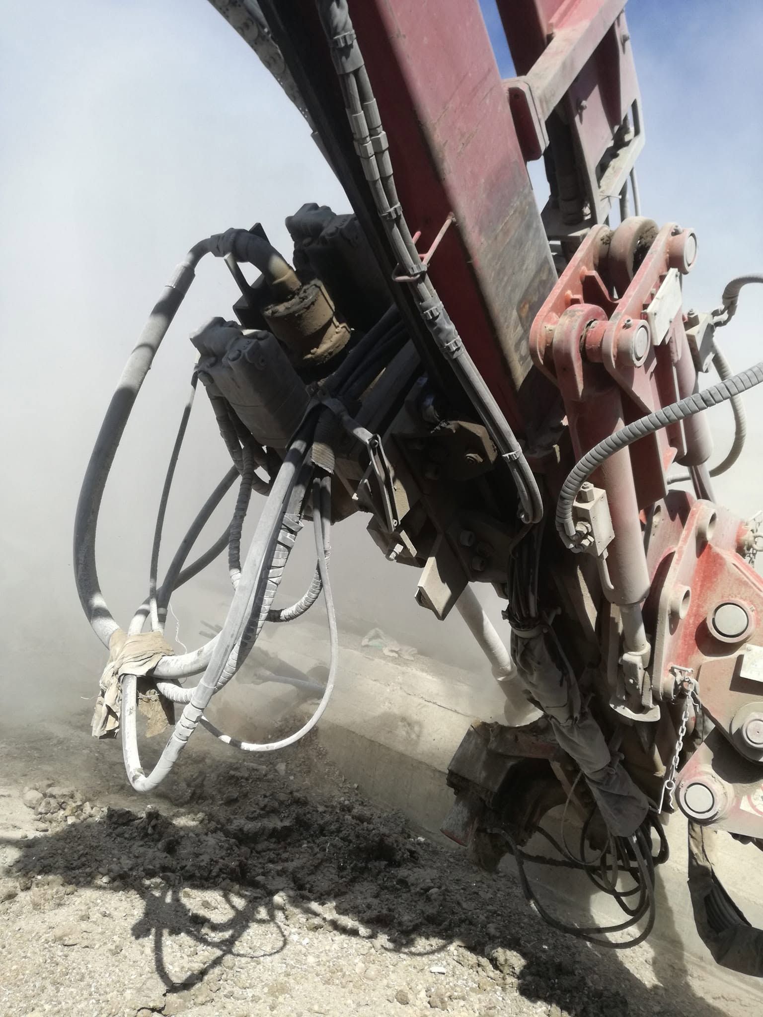 Primo piano dei tubi idraulici e del braccio metallico di una macchina pesante sopra un terreno accidentato, mentre sollevano polvere o detriti.