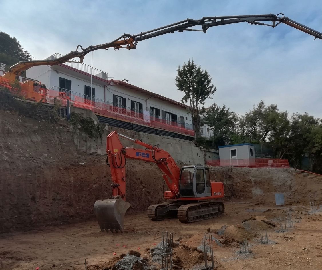 Escavatore e betoniera con pompa in un cantiere in collina accanto a un edificio dal tetto rosso