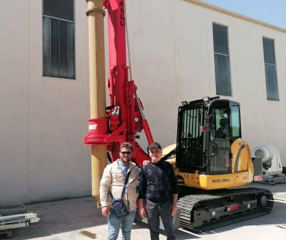 Due persone in piedi accanto a una perforatrice rossa, con un escavatore giallo vicino a un edificio.