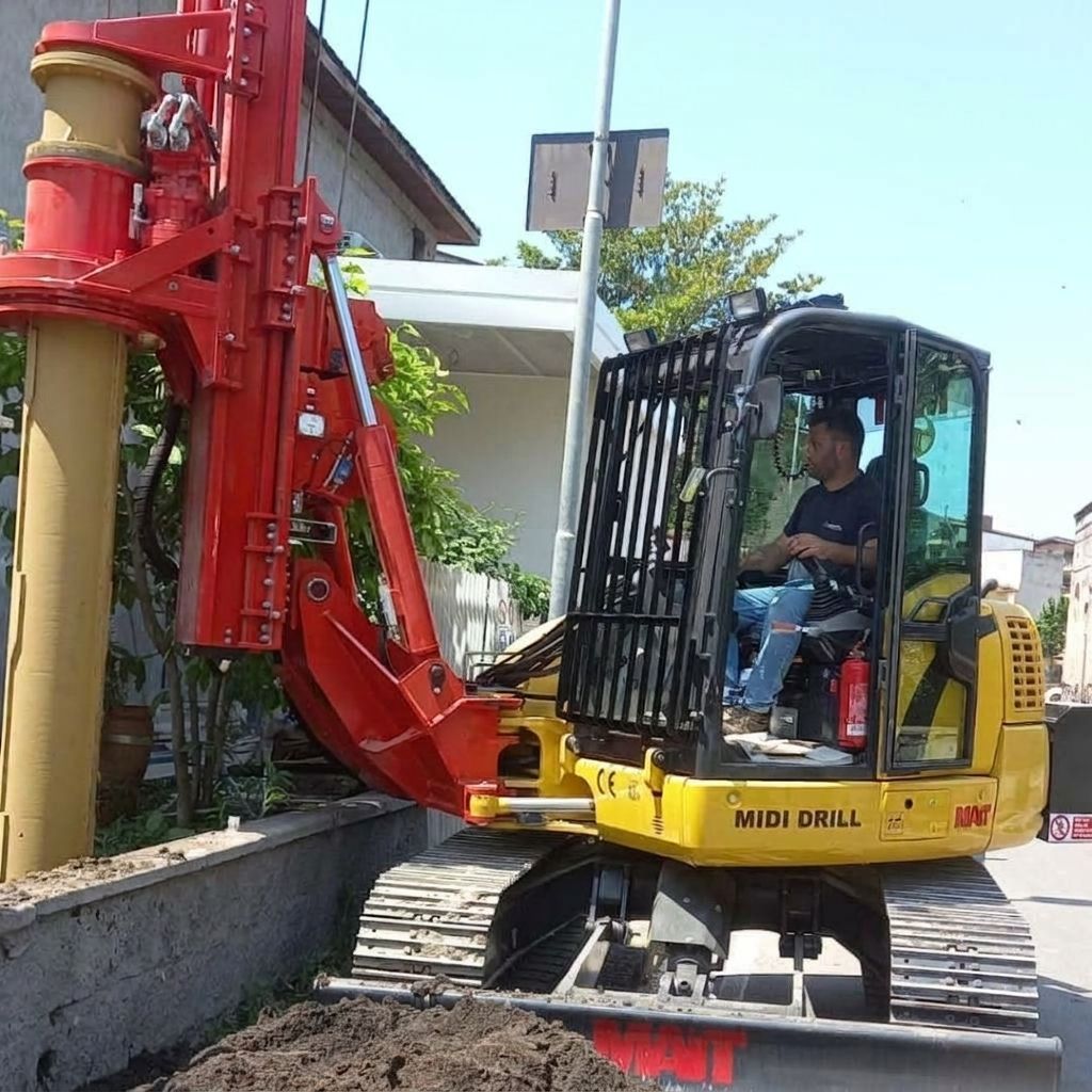 Escavatore rosso e giallo che perfora accanto a un muro di cemento in una strada residenziale, con un operaio nella cabina