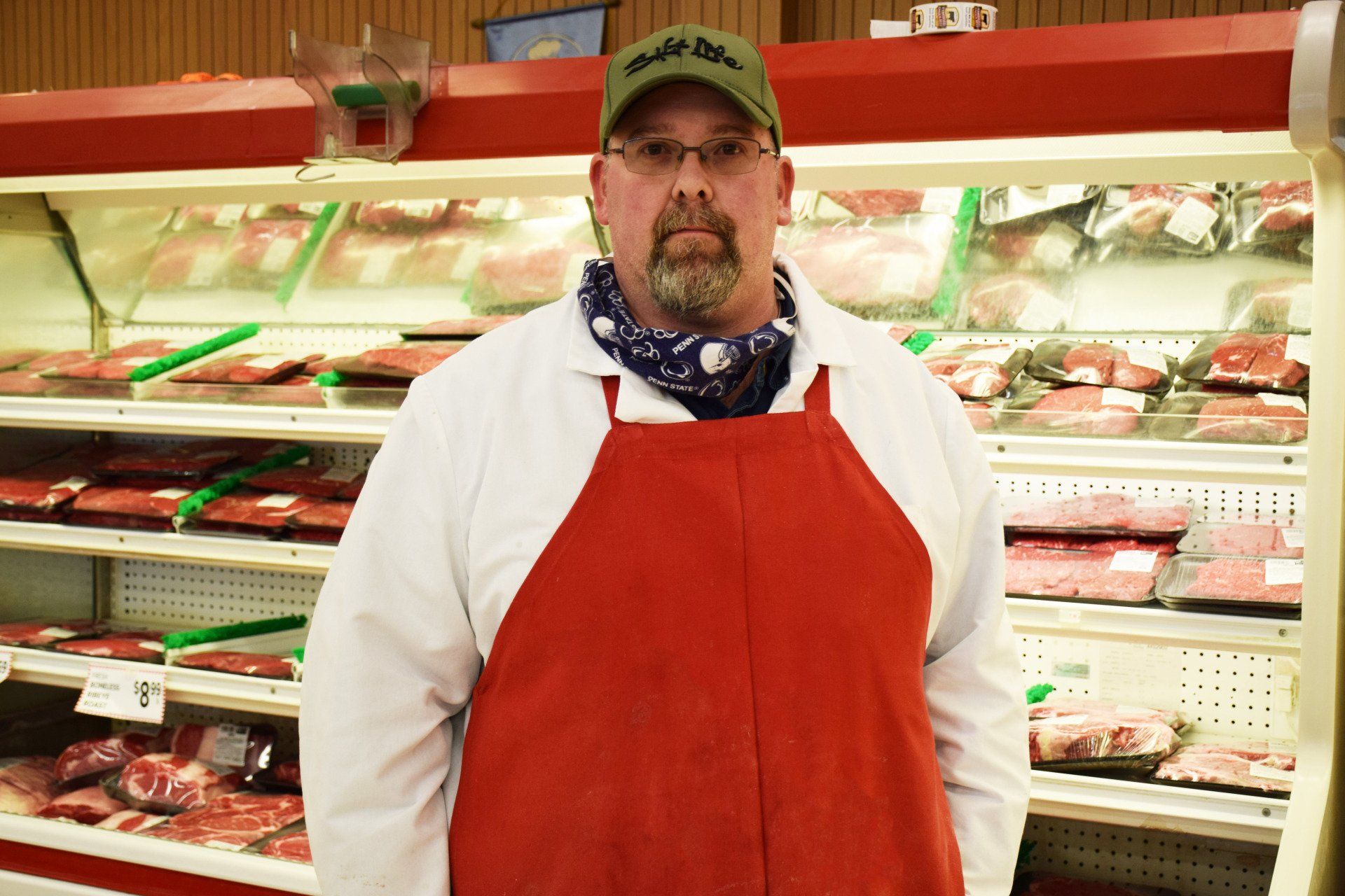 A man in a red apron is standing in front of a meat display case.