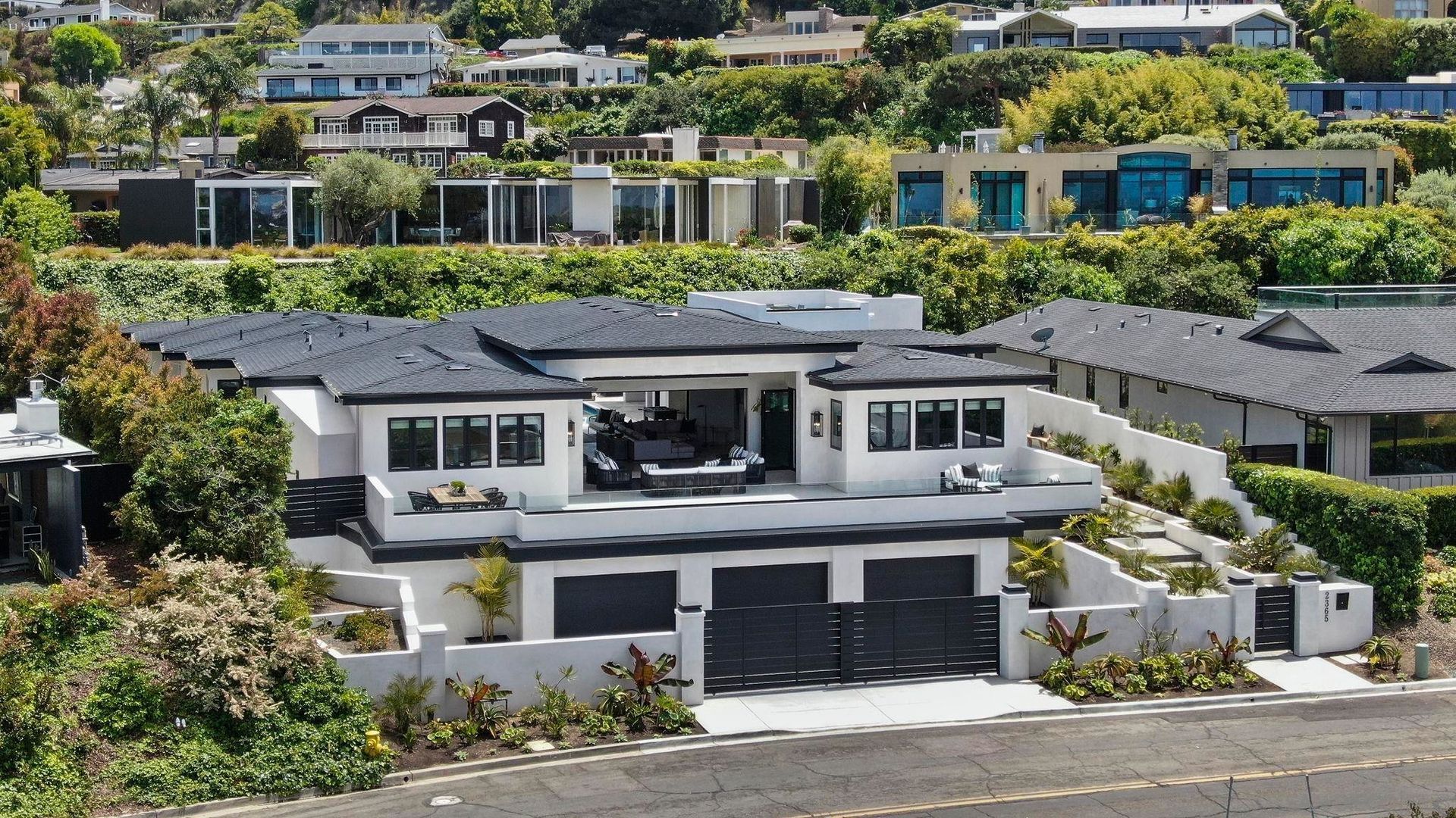 Modern, two-story white house with dark trim and roof, featuring a three-car garage and landscaped hillside setting.