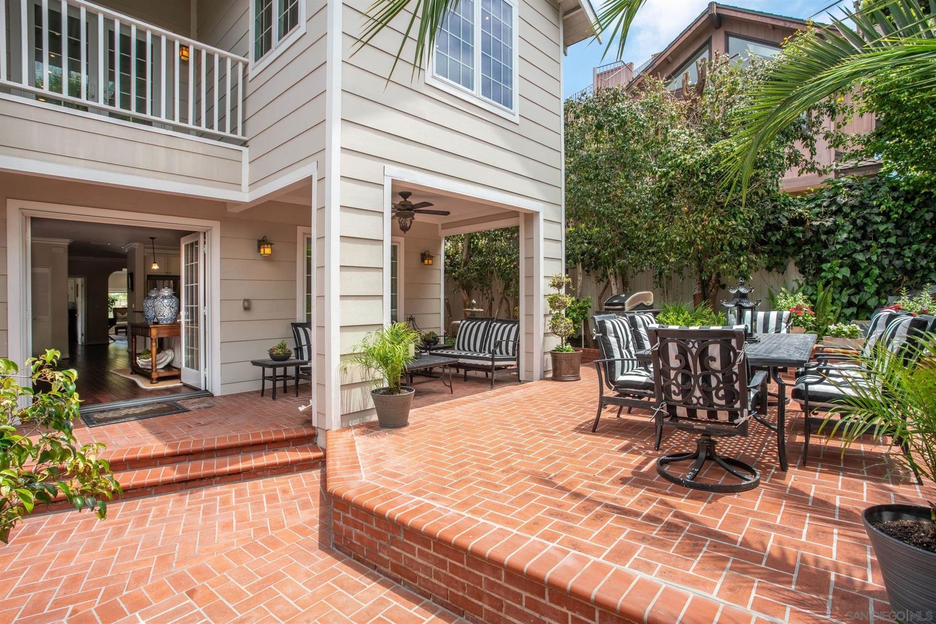 A brick patio with outdoor furniture outside a beige two-story house with a white railing and covered seating area.