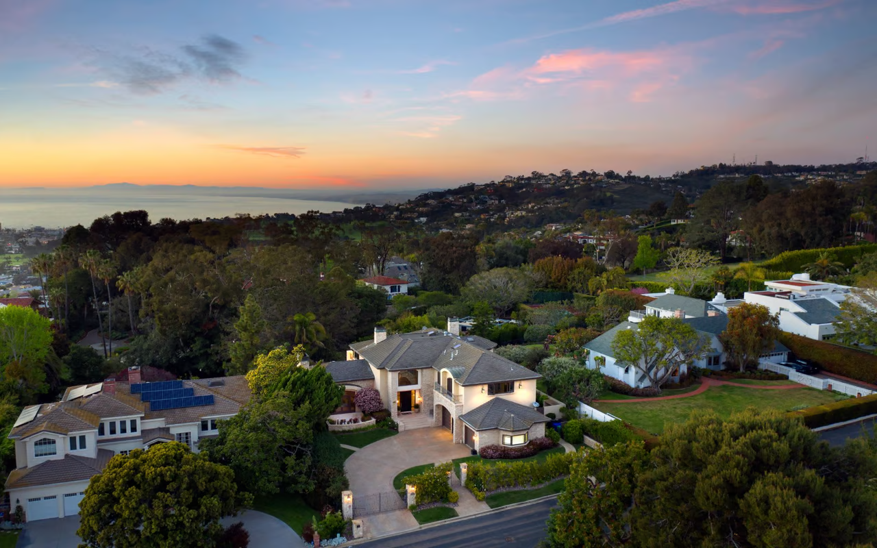 Aerial view of large houses in a hilly residential neighborhood at sunset with a distant view of the ocean.