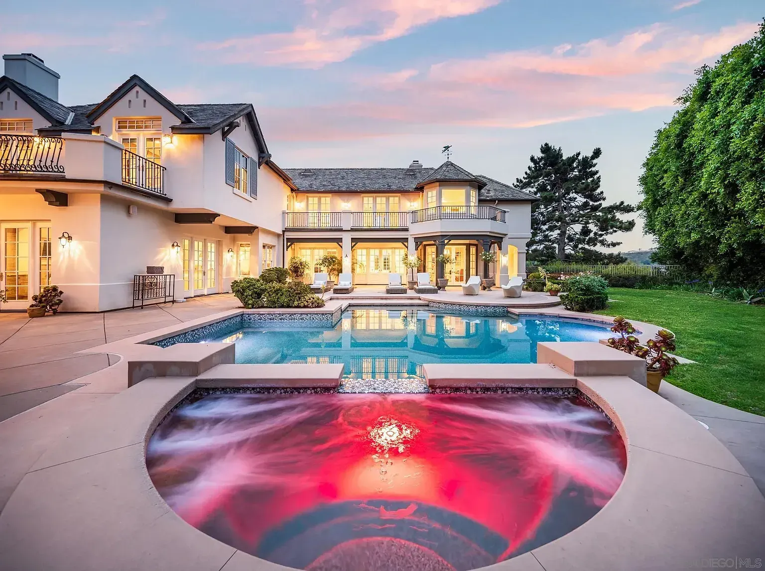 A luxury house with a glowing red hot tub in the foreground, overlooking a swimming pool at sunset.