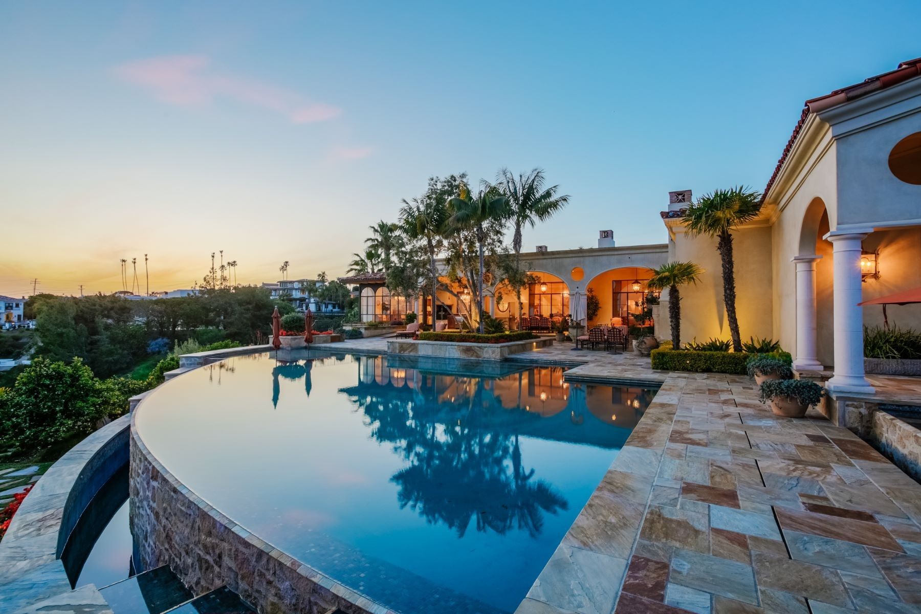 A luxurious infinity pool overlooking a sunset landscape next to a Mediterranean-style home with stone patio and columns.
