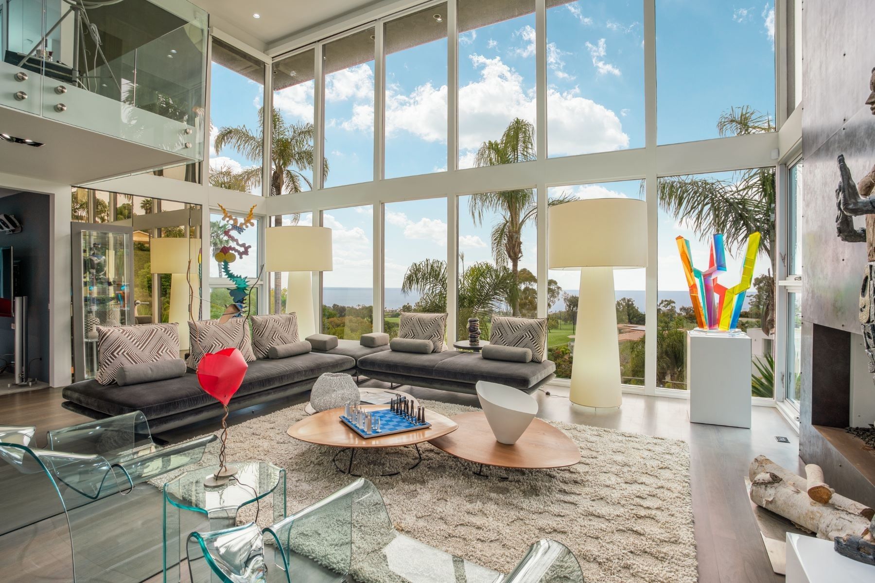Modern high-ceiling living room with a large sofa, glass coffee tables, colorful sculpture, and floor-to-ceiling windows.
