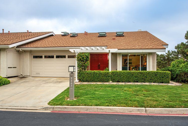 A one-story suburban home with a tan exterior, a brown shingled roof, a garage, and a bright red front door.