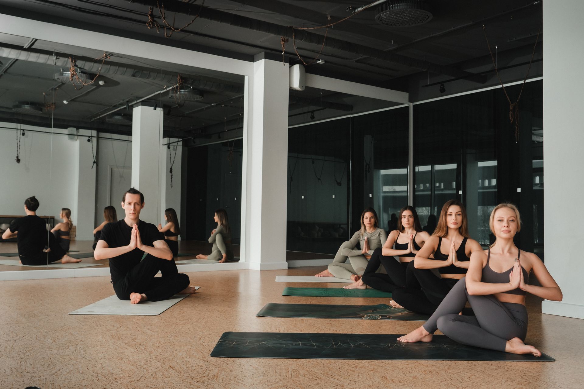 A group of people sits on yoga mats in a bright studio, performing a seated spinal twist pose with palms together.