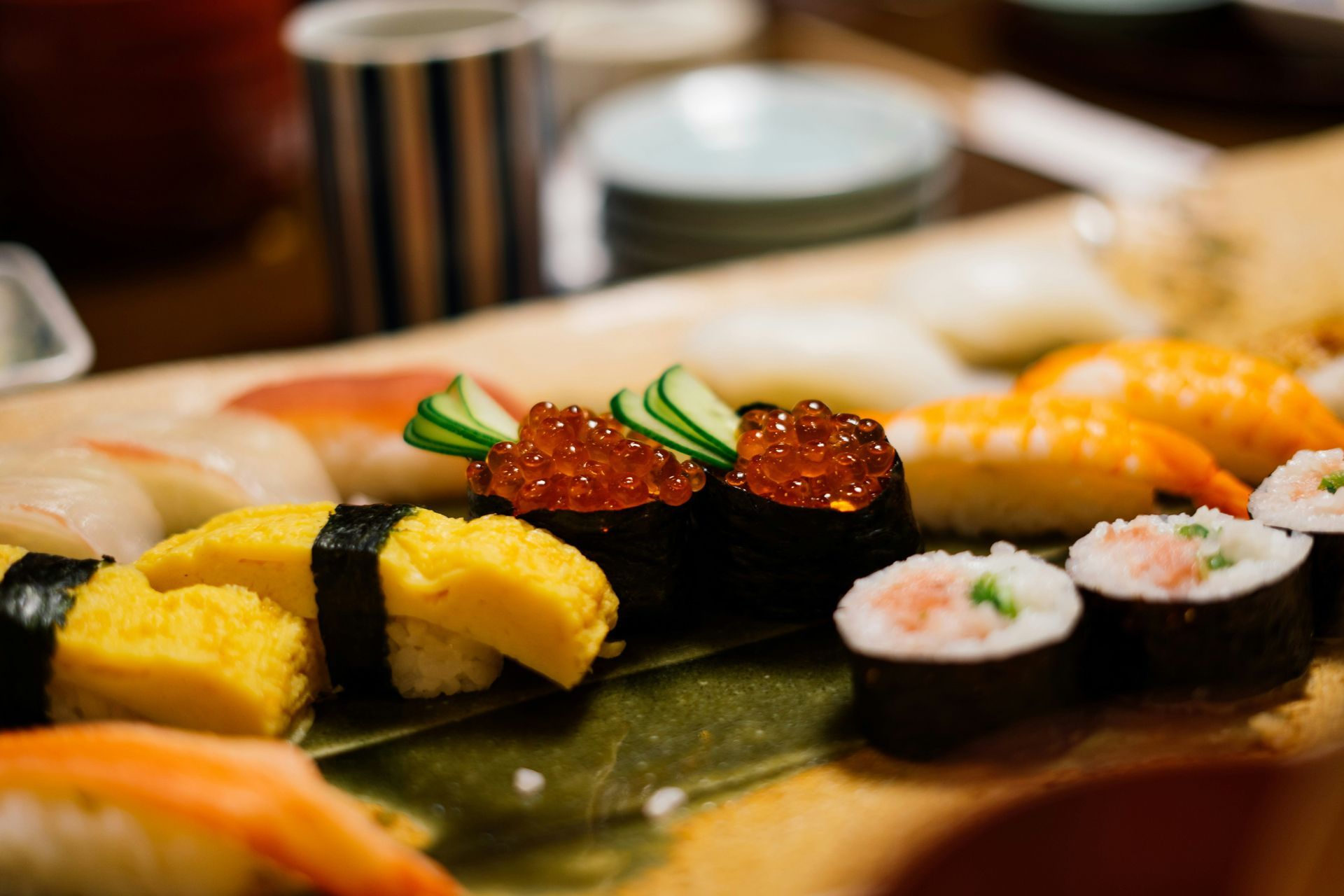 A platter of assorted sushi, featuring nigiri with salmon roe, egg omelet, and small maki rolls on a leaf-lined wooden board.