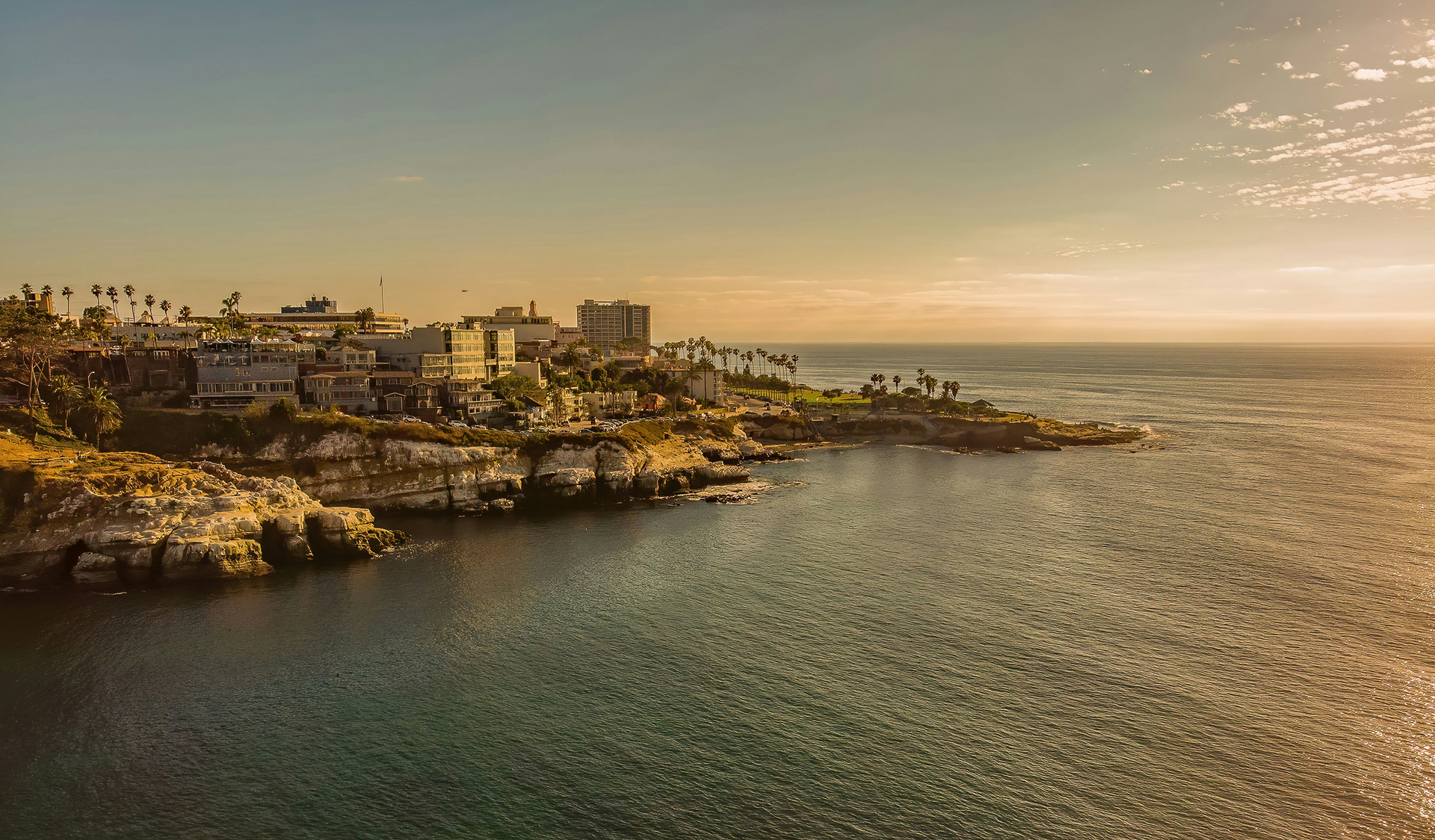 A rocky coastline with buildings overlooks a calm ocean at sunset, bathed in a warm golden light.