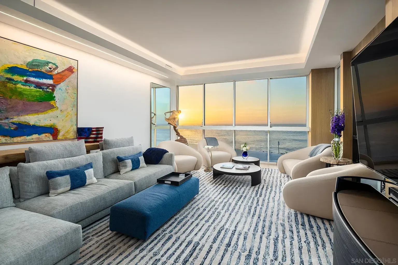 Modern living room with a gray sectional, blue ottoman, plush armchairs, and floor-to-ceiling ocean view at sunset.