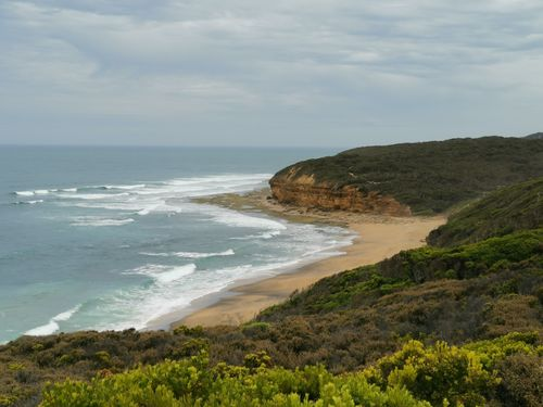 A view of a sandy beach curving along the ocean, bordered by low, rocky cliffs and green coastal vegetation.
