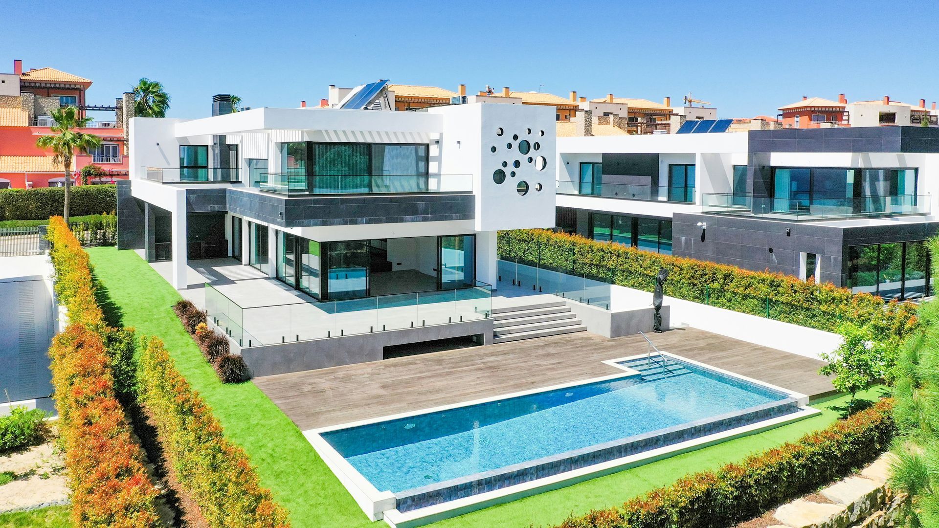 A modern, white two-story villa with large glass windows, a private pool, and green hedging in a sunny, residential area.