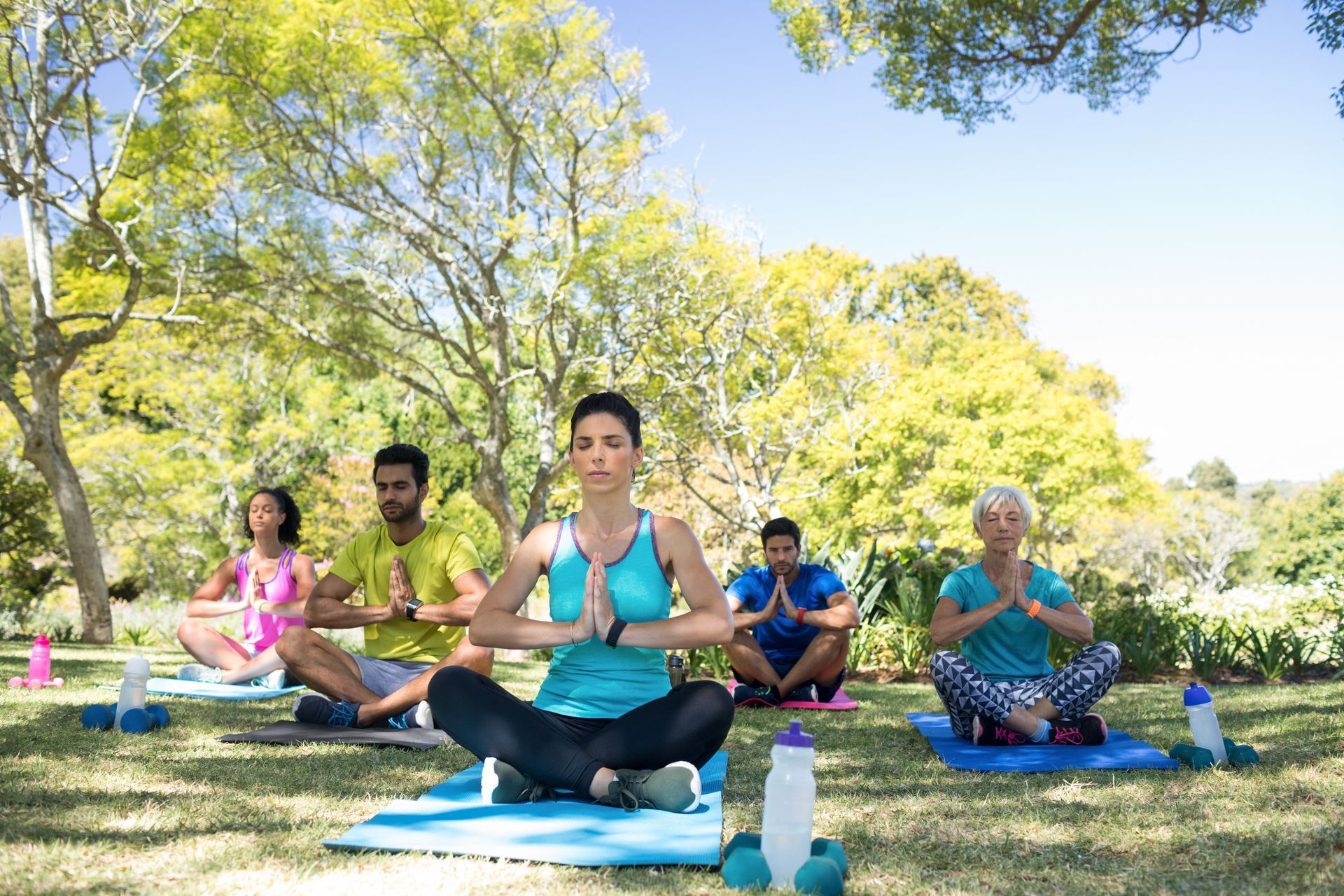 A group of people sits on yoga mats in a park, eyes closed in meditation with hands pressed together in a prayer pose.