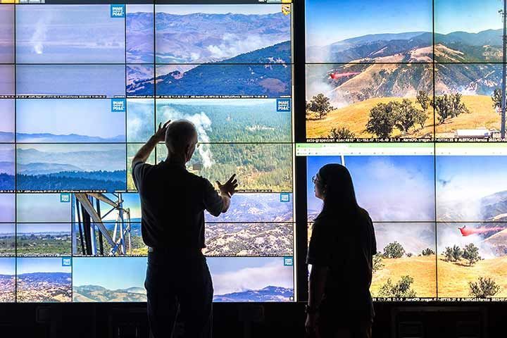 Two people stand before a large multi-screen wall displaying live aerial camera footage of various wildfire incidents.