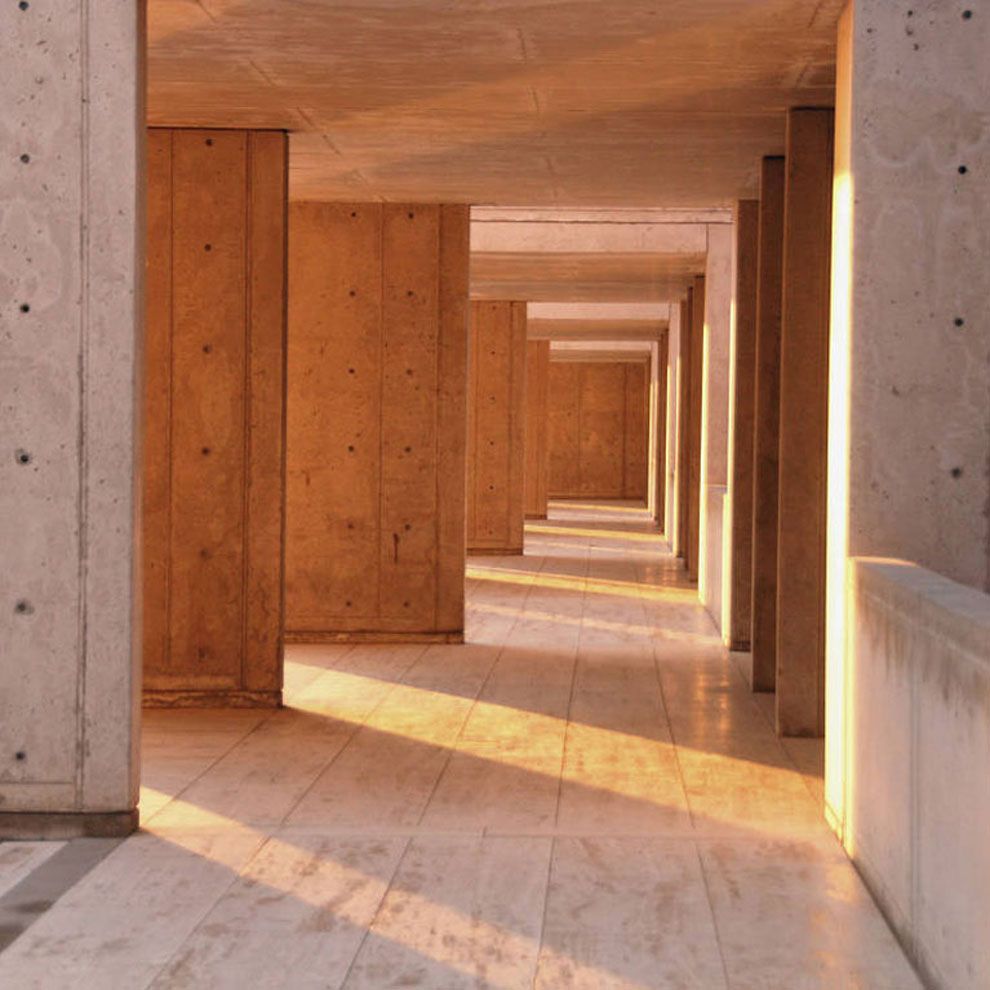 A long corridor with travertine floors and light-colored concrete pillars, bathed in warm, golden afternoon sunlight.