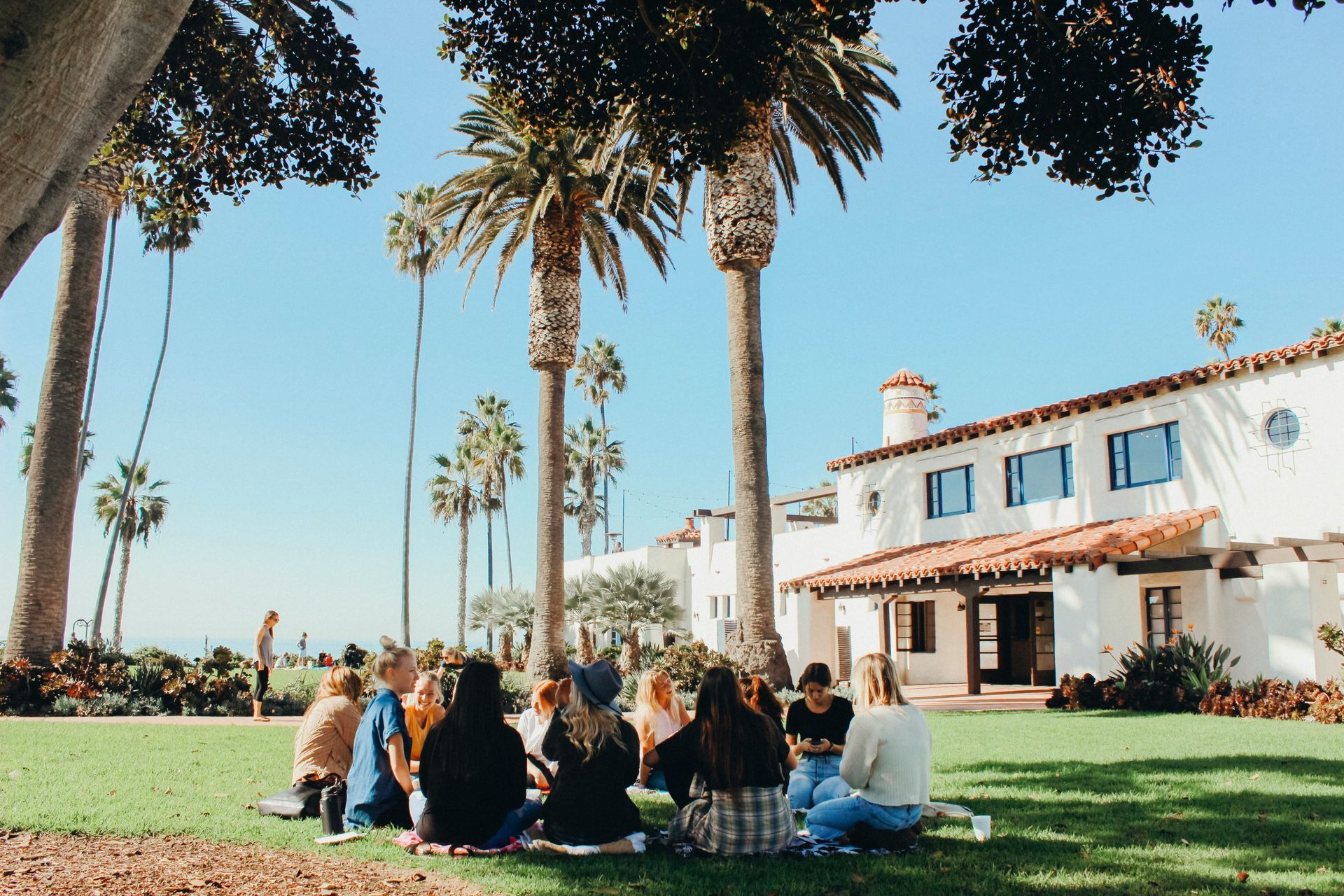 A diverse group of people sitting in a circle on a green lawn in front of a white Spanish-style building with palm trees.