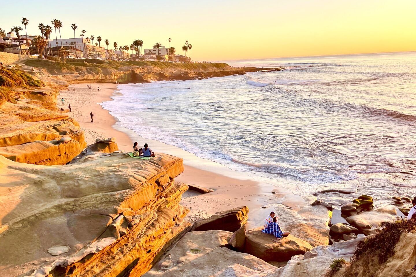 People sit on golden cliffs overlooking a sandy beach and ocean waves during a warm, sunset-lit day.