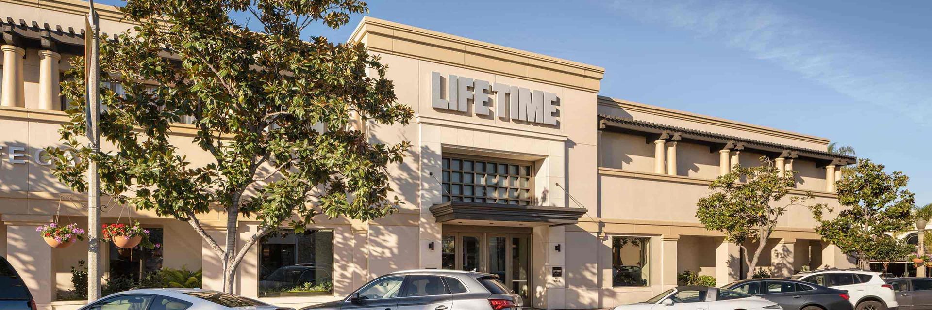 A tan-colored Lifetime gym building facade with large trees in front and a parking lot with cars below.