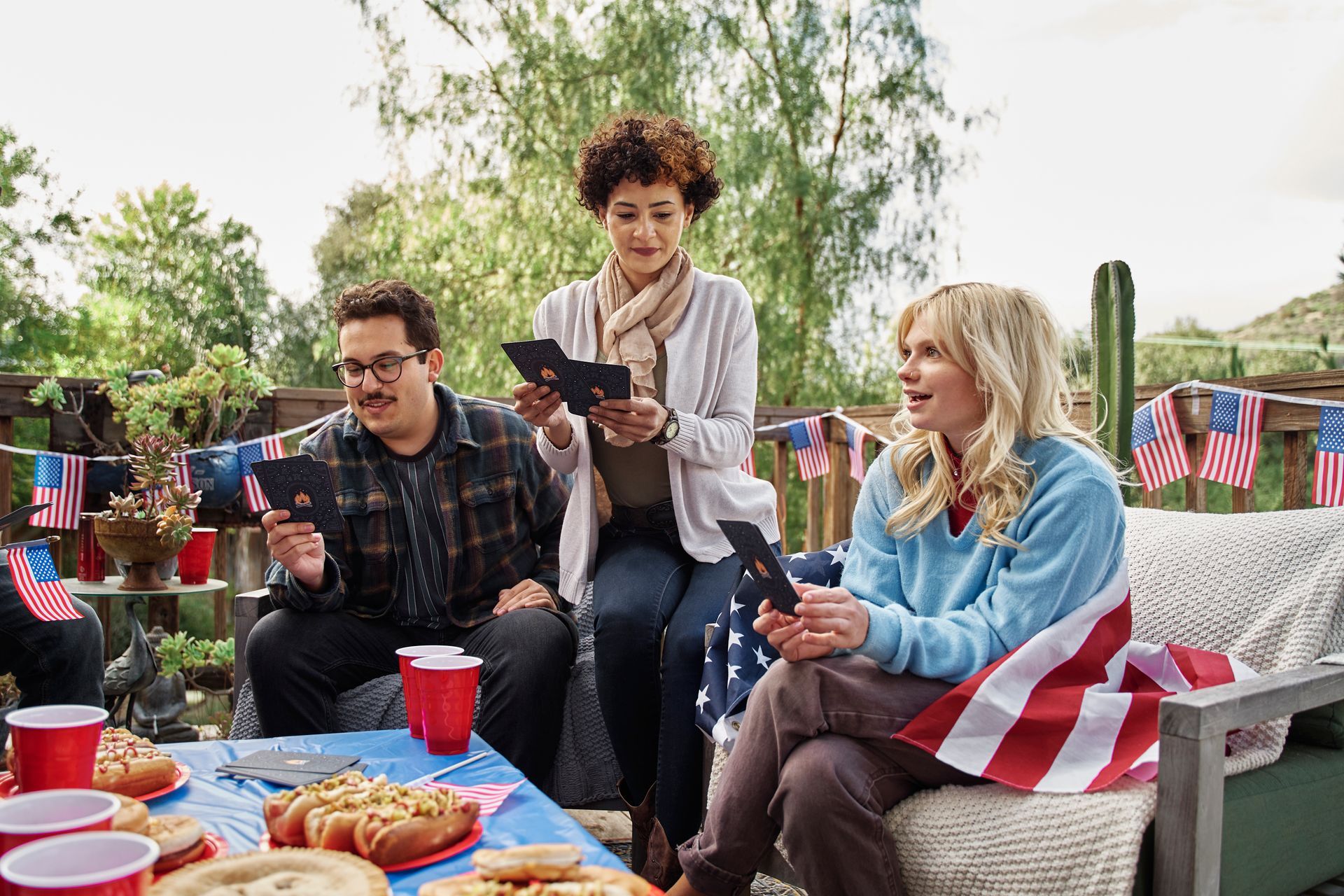 Three people play cards around a table with snacks and American flag decorations on an outdoor deck.