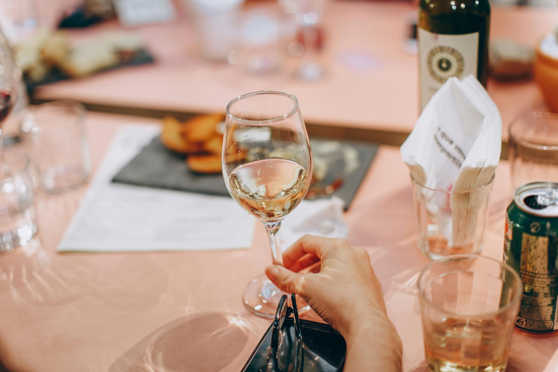 A person holding a glass of white wine at a table set with food, drinks, and a bottle on a pink tablecloth.
