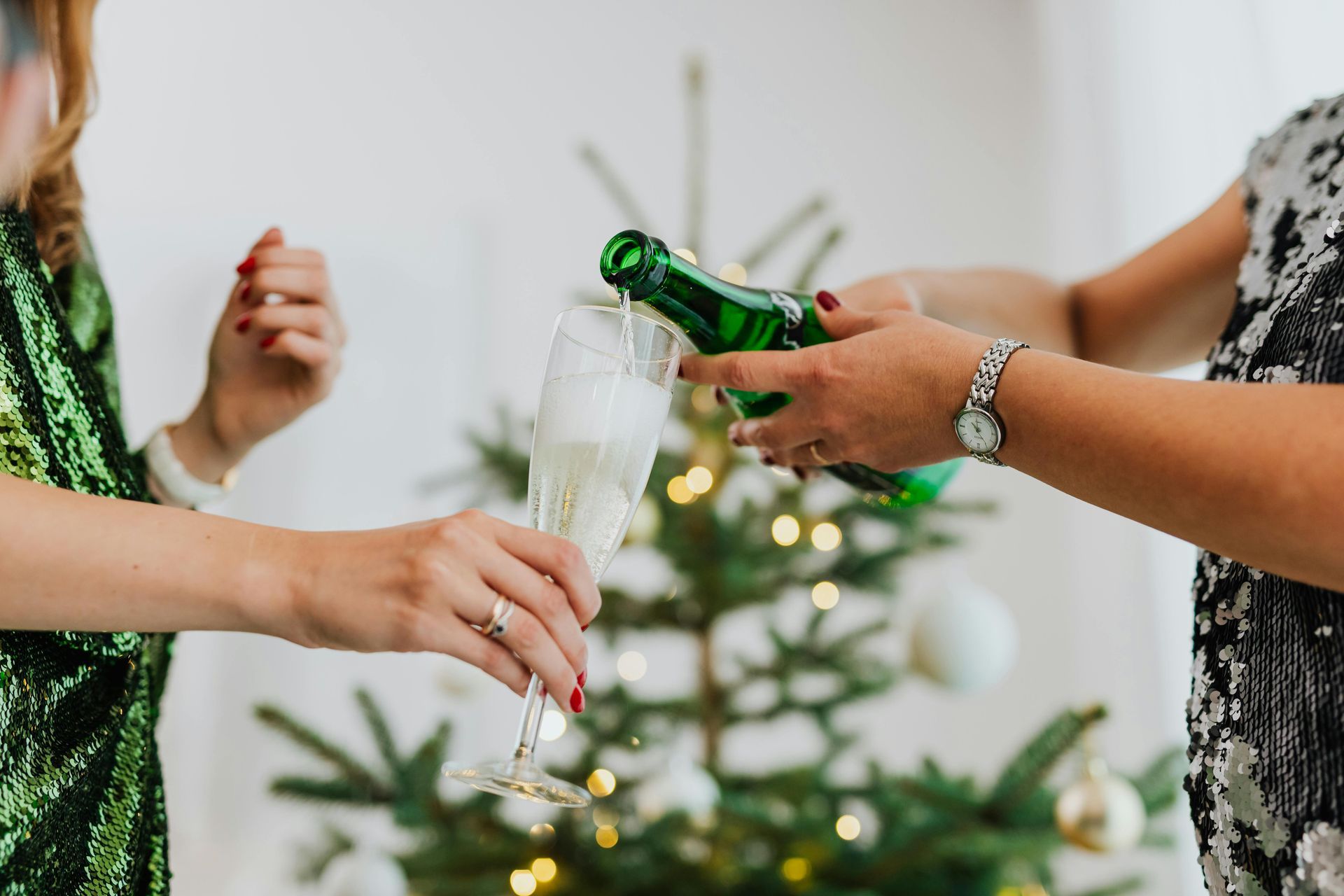 Someone pours sparkling wine from a green bottle into a glass held by another person in front of a decorated Christmas tree.