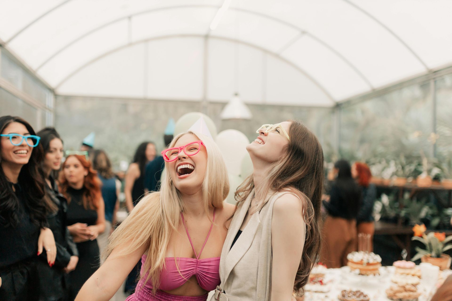 Two laughing people wearing party glasses at an indoor gathering, with blurred guests and dessert tables in the background.