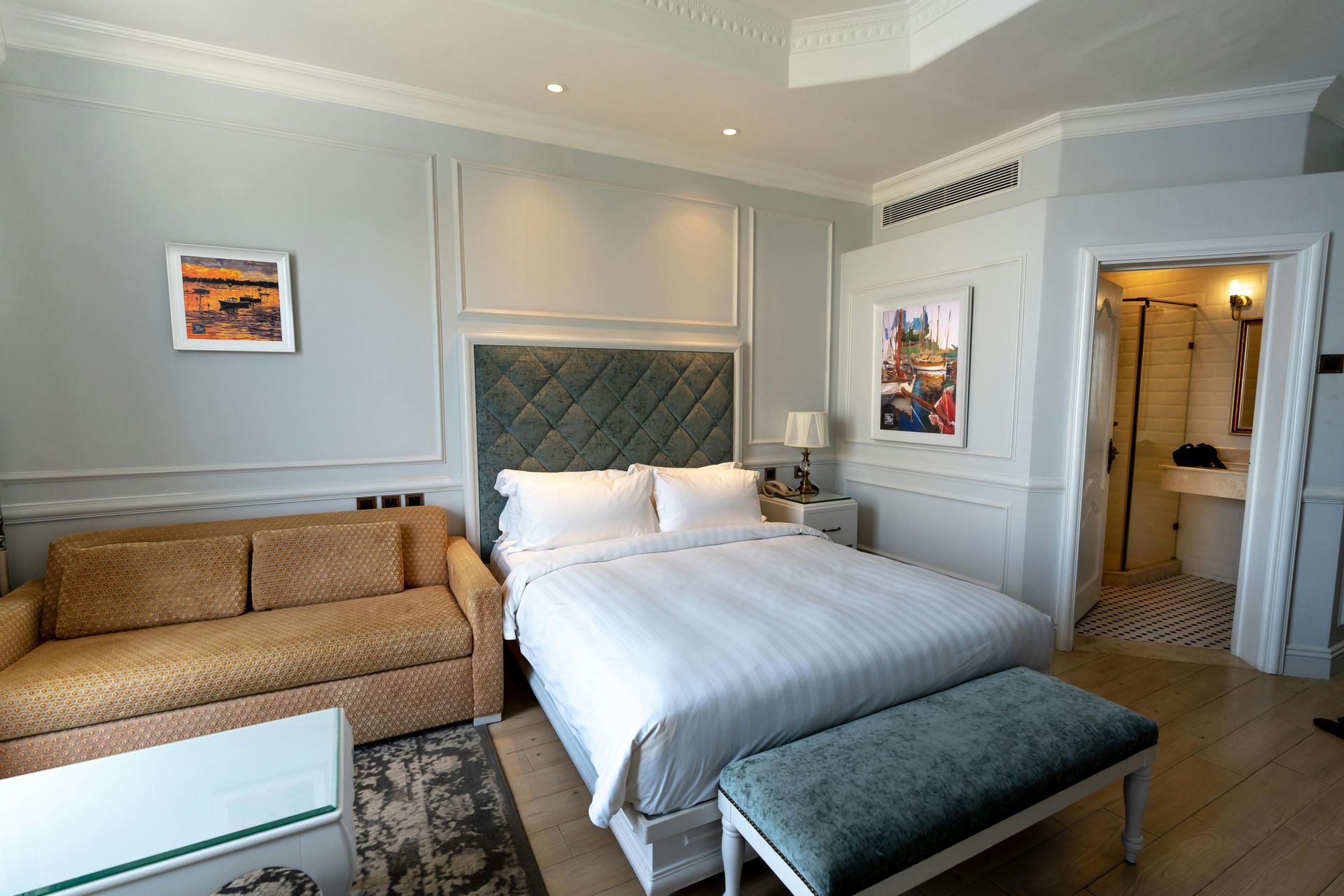 A hotel room featuring a bed with white bedding, a gold sofa, a glass coffee table, and an adjacent bathroom.