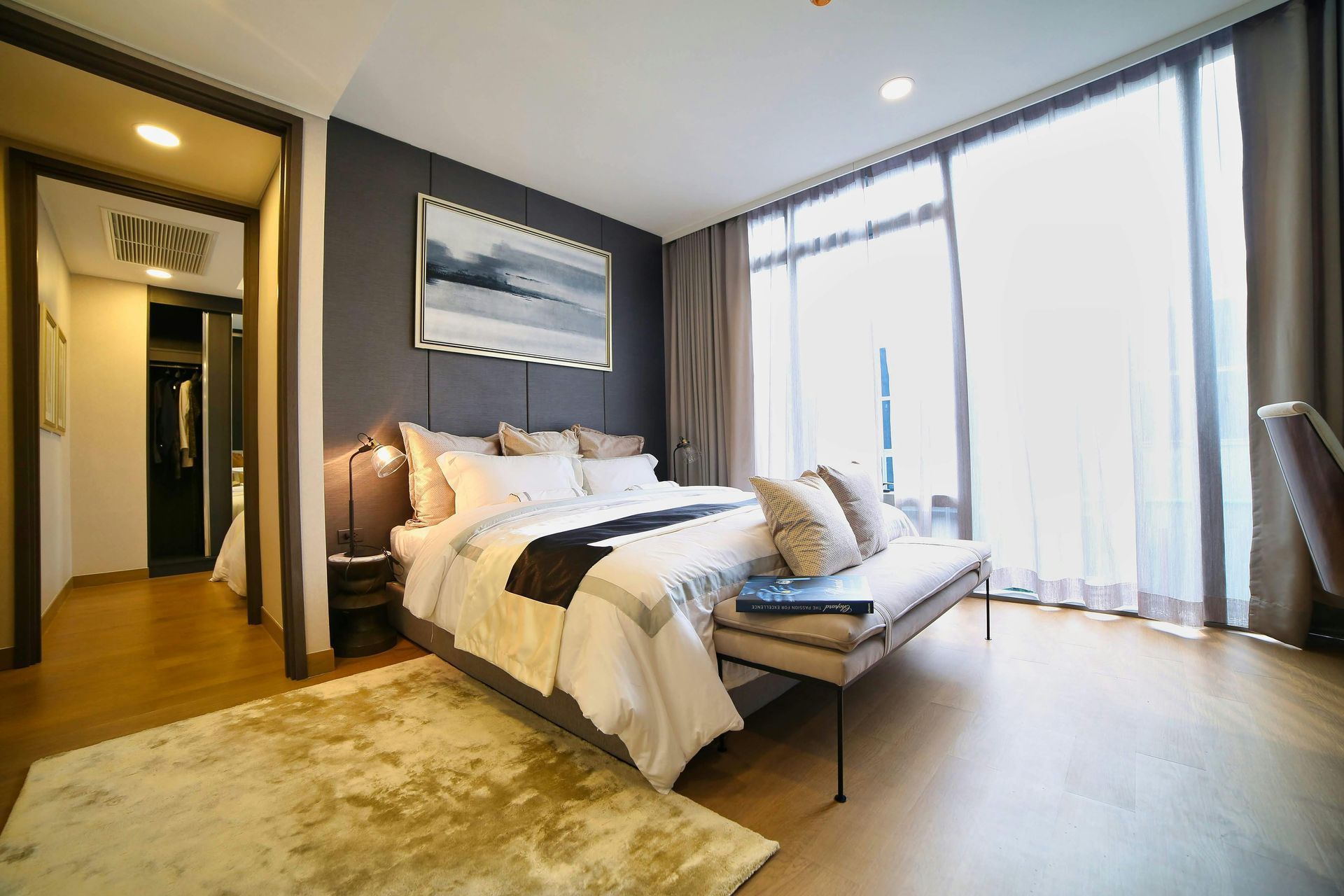 Modern bedroom with a gray accent wall, king bed with white bedding, a bench, and floor-to-ceiling windows with curtains.