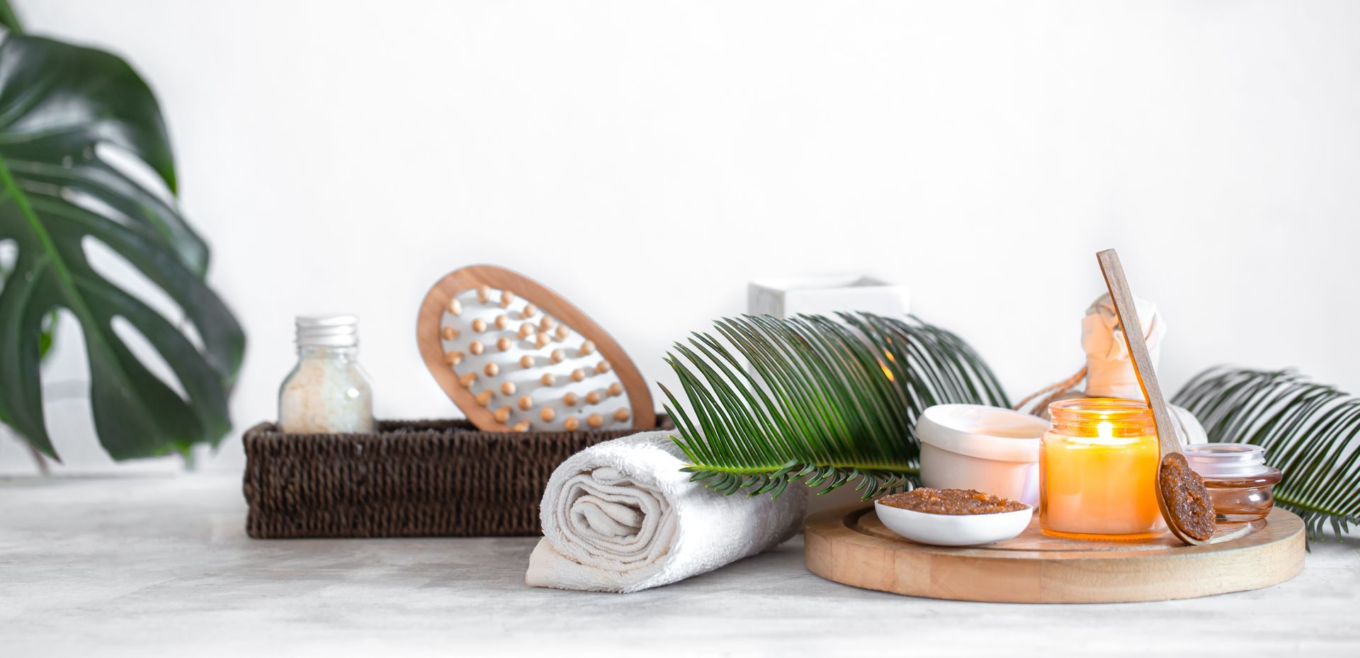Spa setting with massage tools, a white towel, a lit candle, and green leaves on a light surface.