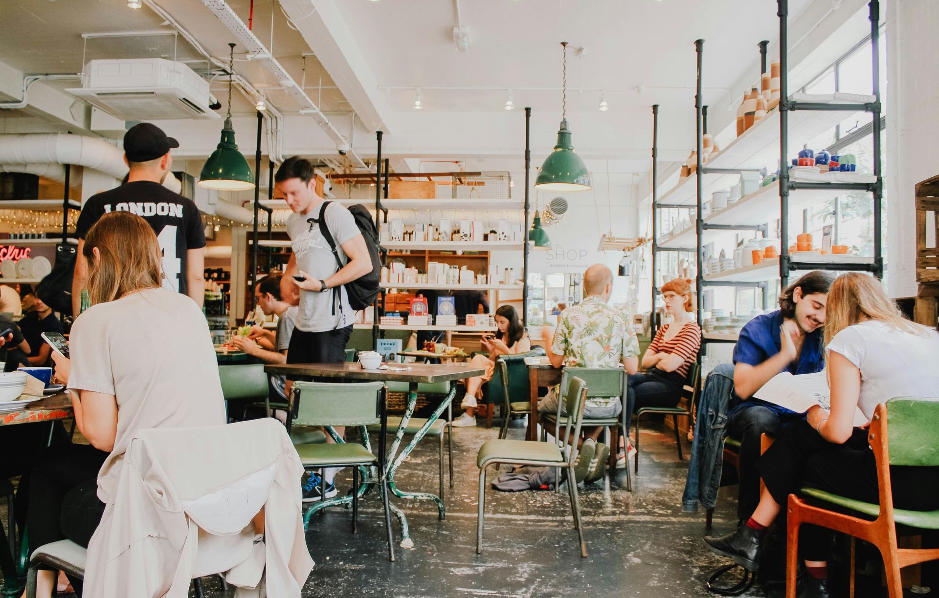 People sit at tables and work in a brightly lit, modern café with open shelving and industrial-style green pendant lights.