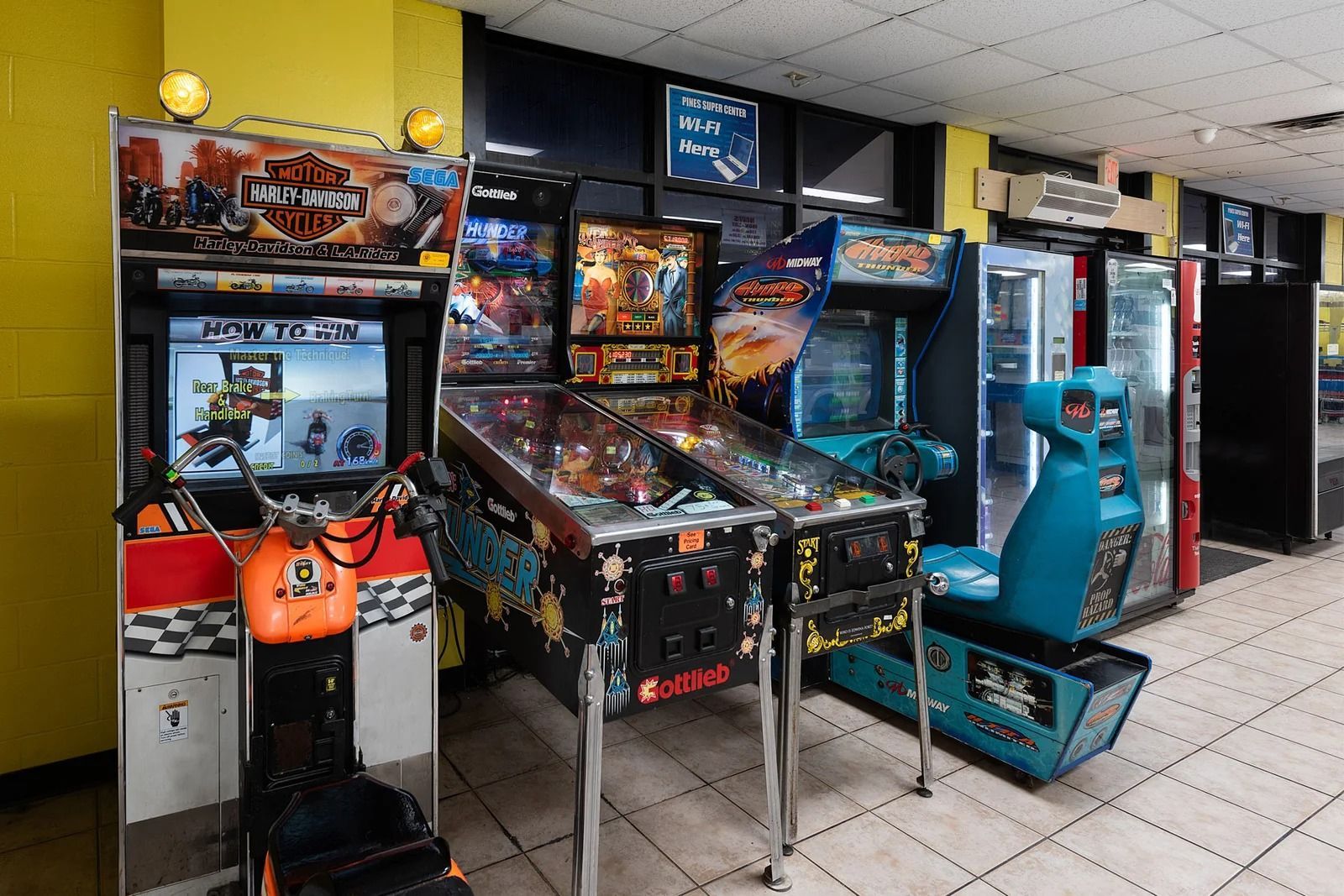 Arcade games, including pinball machines and motorcycle racing game, in a brightly lit room.