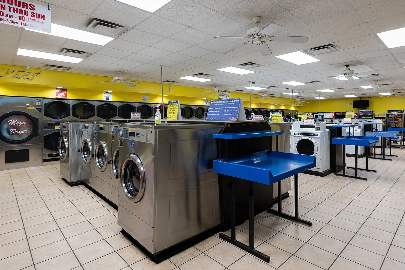 Interior of a laundromat with rows of washers and dryers; yellow walls, white ceiling, and tiled floor.