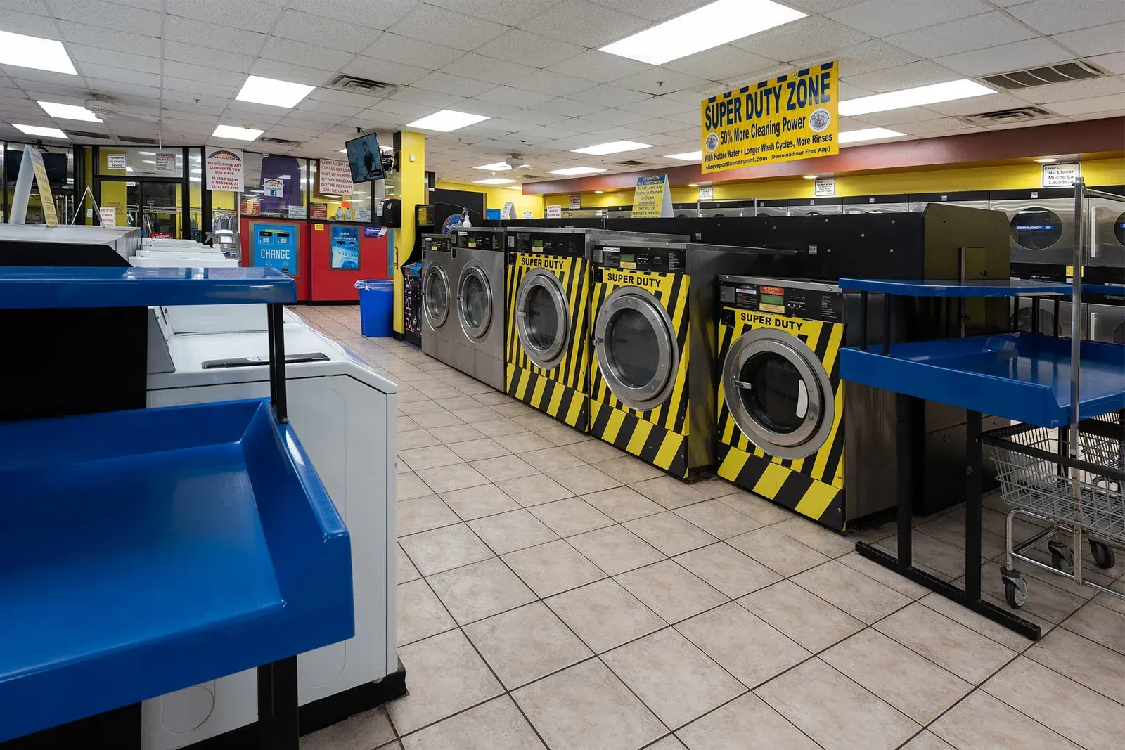 Interior of a laundromat with rows of washers and dryers. Yellow and black striped washing machine, blue surfaces, and tiled floor.