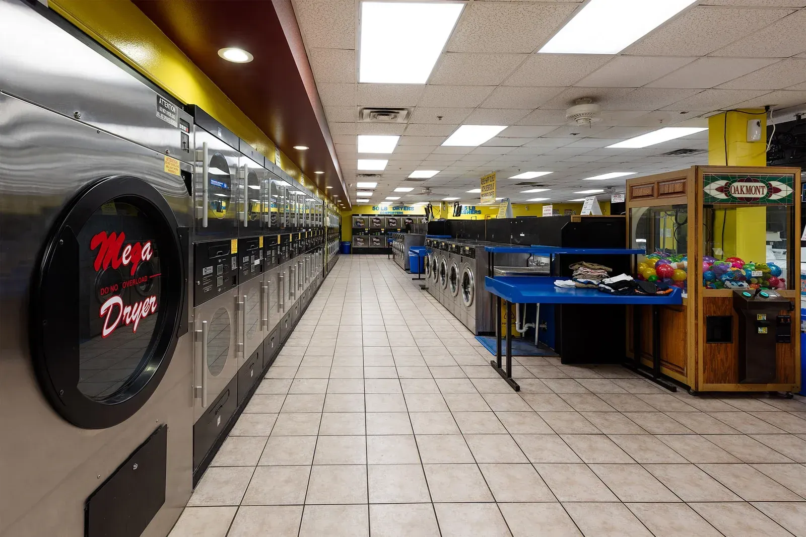 A laundromat with rows of washers and dryers, a blue table, and an arcade game.