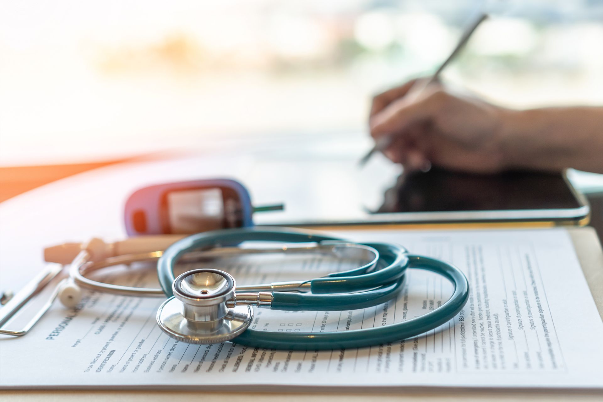 A Stethoscope is Sitting on Top of a Clipboard Next to a Person Writing on a Tablet