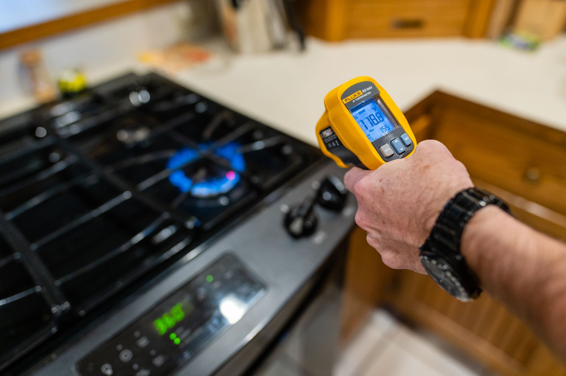 Hand holding a yellow infrared thermometer aimed at a lit gas stove burner in a kitchen