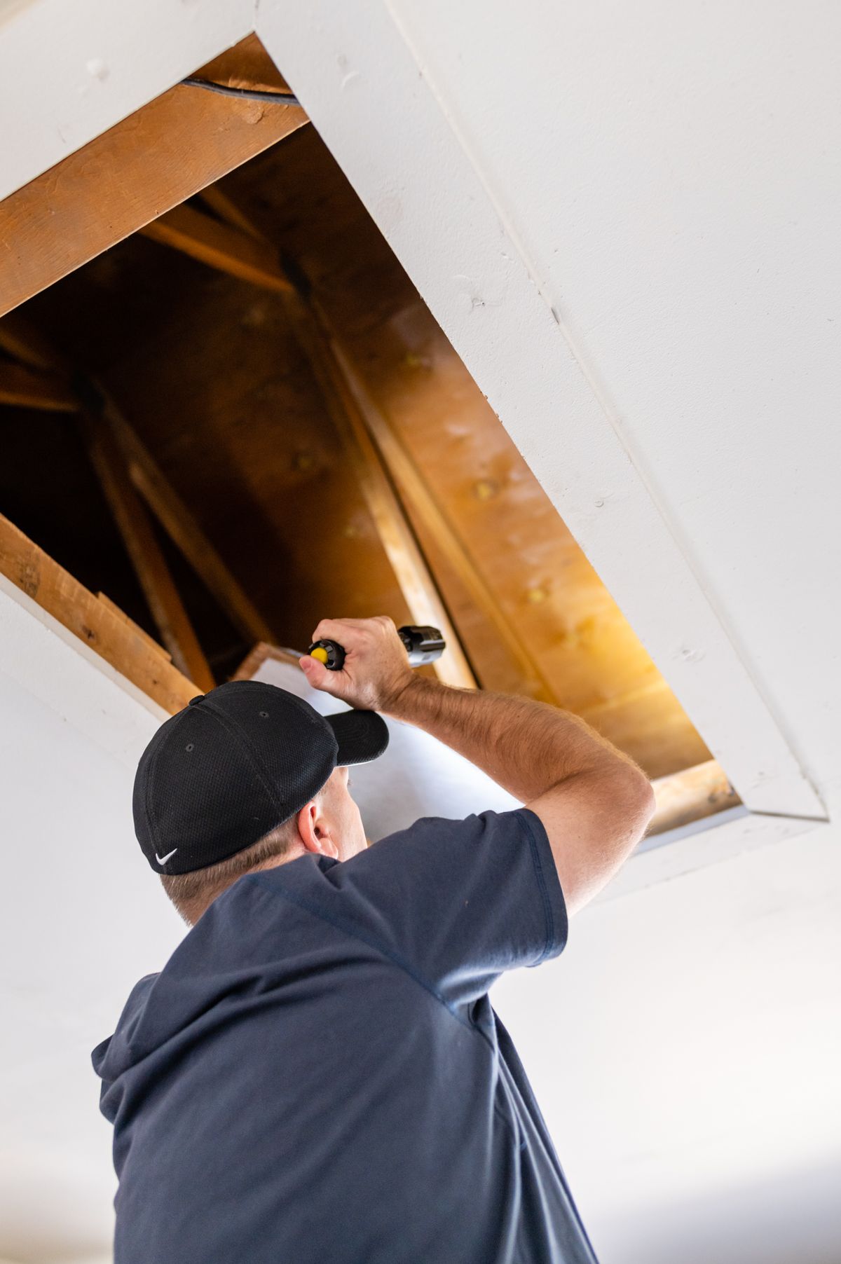 Man in a cap using a flashlight to inspect an attic opening in the ceiling