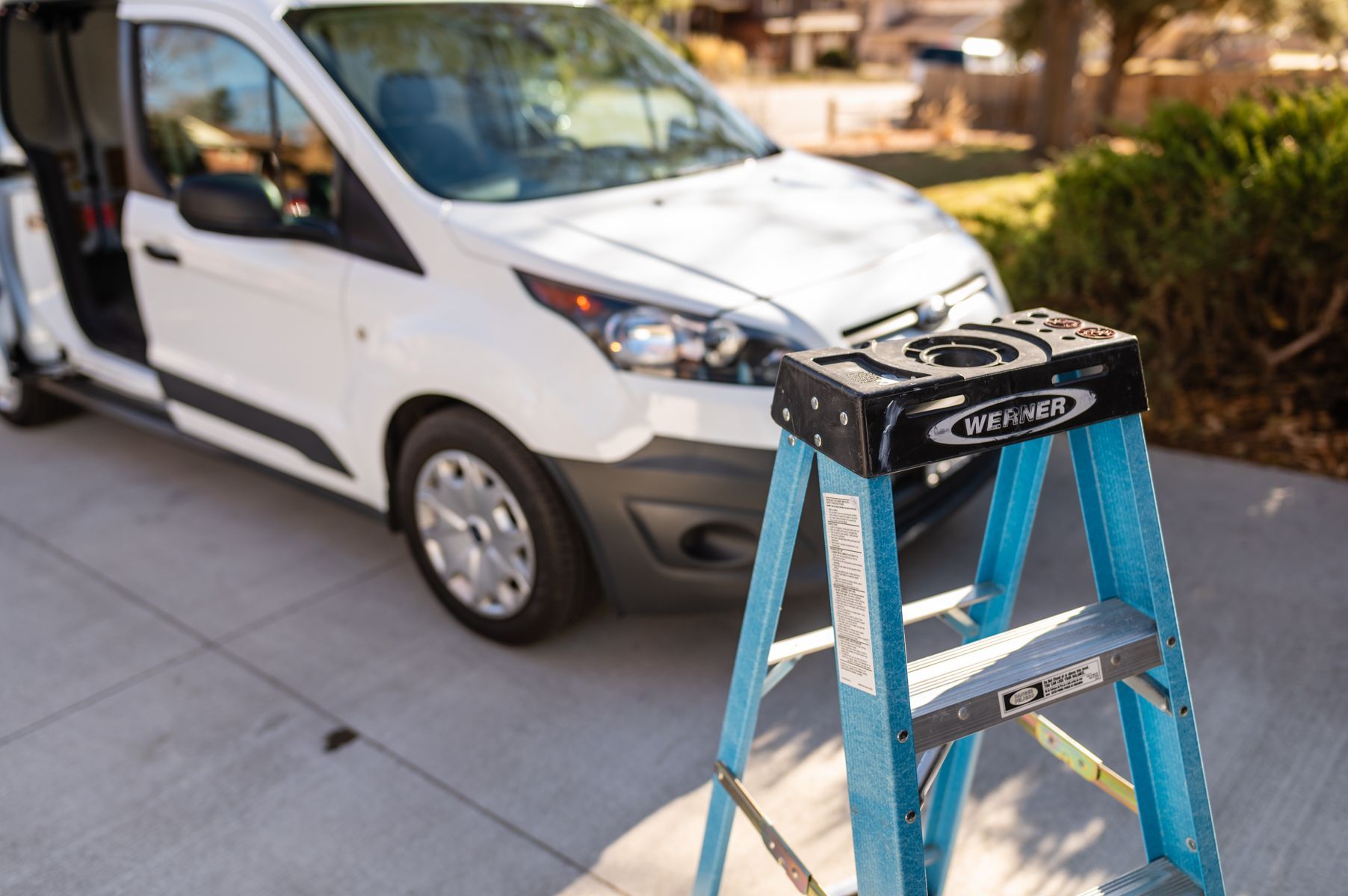 Blue step ladder in front of a white van parked on a driveway with a garage in the background