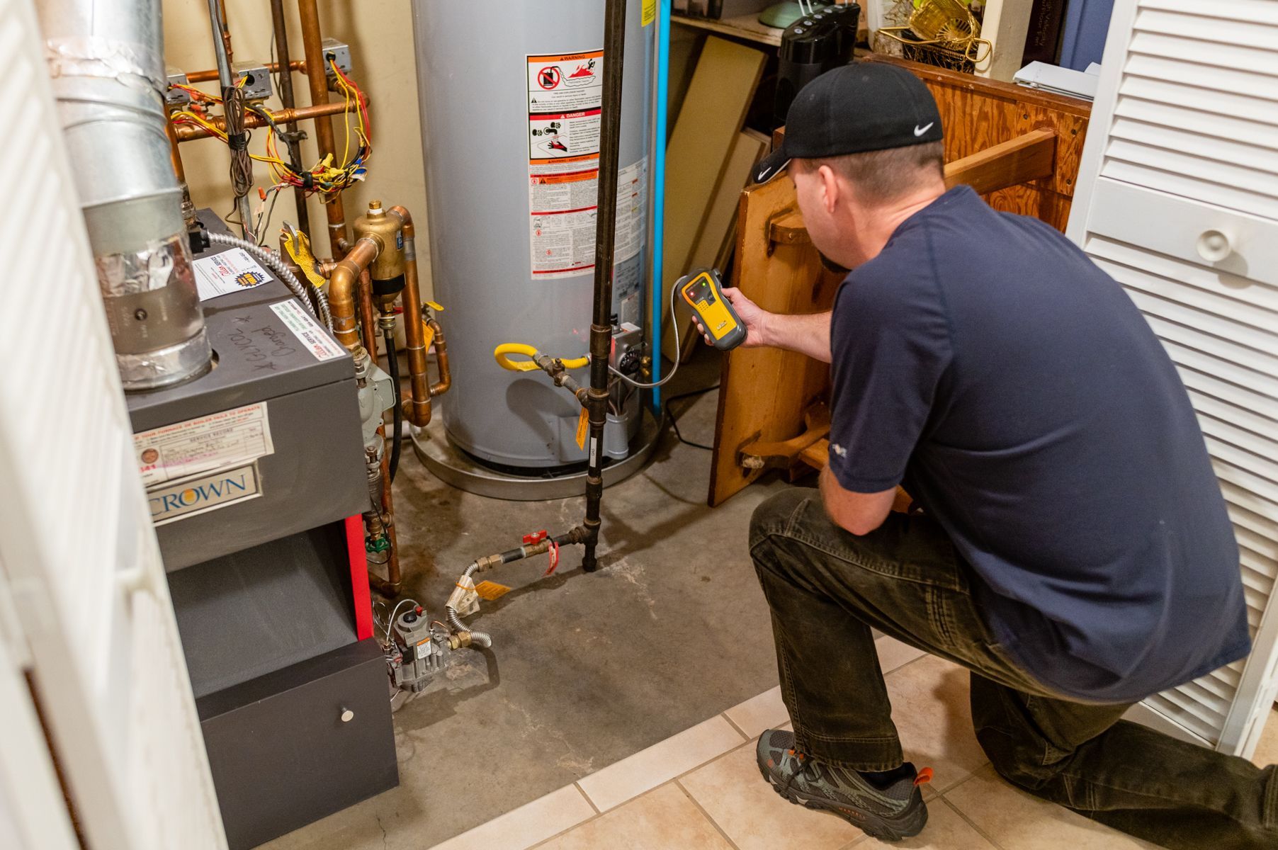 Technician kneeling by a furnace, inspecting pipes and wiring in a utility room.