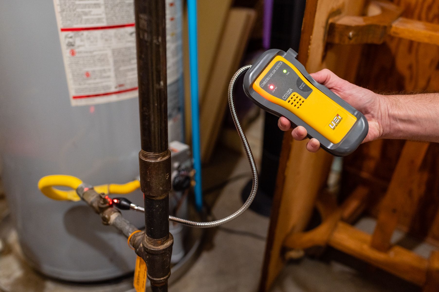 Technician using a handheld gas leak detector on a pipe next to a water heater.