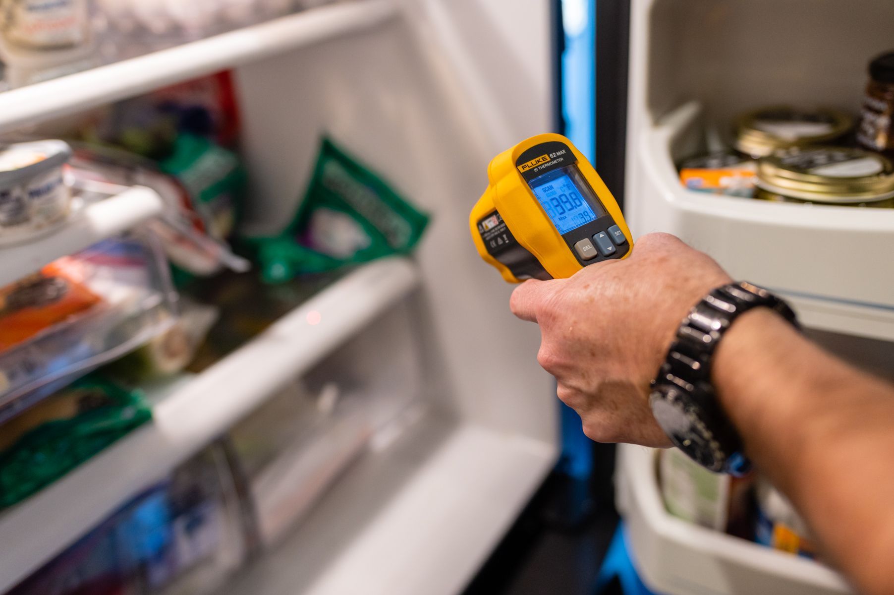 Hand holding a yellow infrared thermometer aimed at items inside an open refrigerator