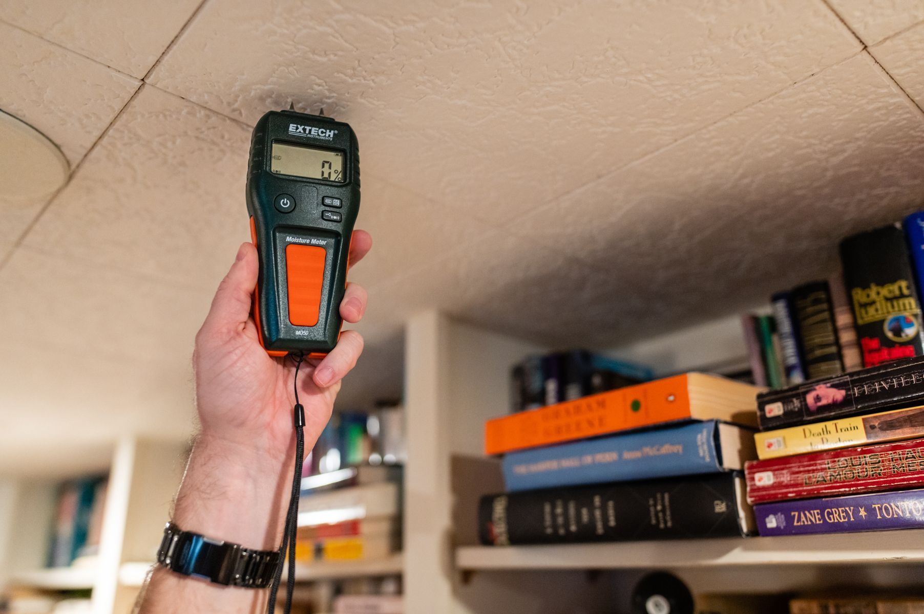 Handheld orange and black device raised toward a ceiling in a room with shelves of books and boxes