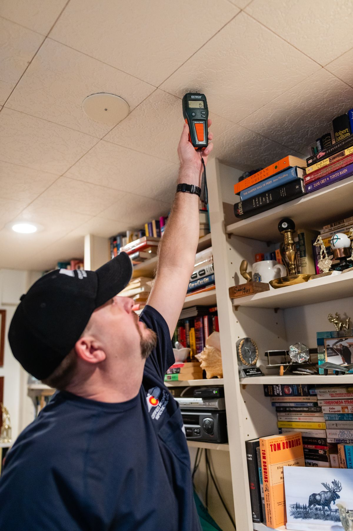 Person in a navy shirt and black cap reaching up with a handheld meter toward the ceiling in a room with shelves.